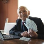 Happy senior businessman showing cash at his desk with a laptop, indoors.